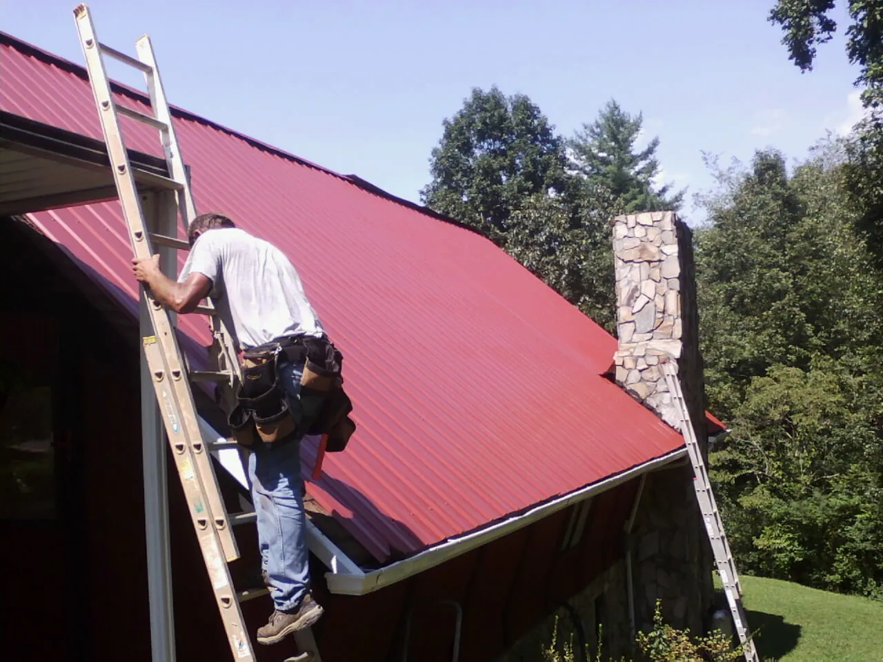 Roofer on a ladder beside a red metal roof and stone chimney — Soffit & Fascia Repair by Roofing Brothers in South Orange